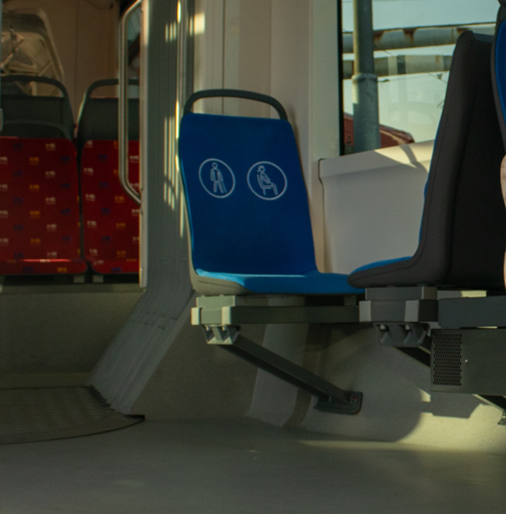 Na obrázku je záber na sedačku v autobuse, ktorá je modrej farby a na chrbtovej časti má výrazné biele piktogramy s označením človeka s bielou palicou či nastávajúcej matky.