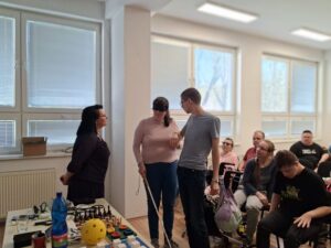 Dvaja klienti muž a žena stoja pri sebe. Žena má na očiach zatemňovaciu masku a muž ju drží za ruku. Vedúca Krajského strediska pani Sváková obom vysvetľuje techniku vedenia nevidiaceho. Ostatní klienti sedia sa pozerajú. 