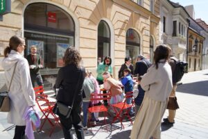 Skupinová fotka, na ktorej sú stojací ľudia okolo stolov s prezentačnými vecami ÚNSS. Vedúca Krajského strediska drží mikrofón v ruke a hovorí o sociálnej práci. 