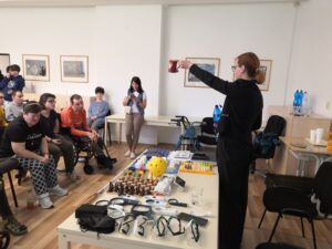 Sociálna pracovníčka stojí za prezentačným stolom, v ruke drží pohár so zdvihnutou rukou, do ktorej naliala vodu z fľaše. Klienti sedia a pozorne ju sledujú. 