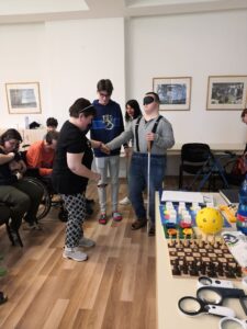 Dievča podáva ruku chlapcovi s bielou palicou, aby mu pomohla. Pri nich stojí mladý muž spolu so sociálnou pracovníčkou DSS ROSA. Túto scénu sledujú sediaci klienti na stoličkách. 