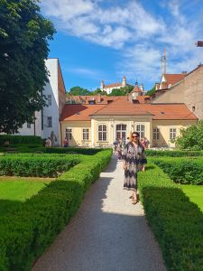 Záhrada obkolesená malými kríkmi v pravidelných tvaroch znázorňujúc úzke uličky. V strede stojí mladá dáma v tmavých dlhých šatách, ktorá sa usmieva. Svieti slnko a je teplo. 