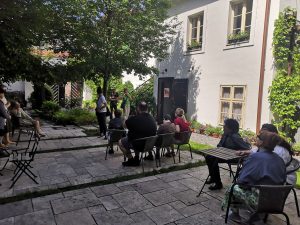 Veľká priestranná záhrada, v strede je betónová podlaha, na ktorej sú postavené stoly a stoličky a ľudia sediaci a počúvajúci  výklad o histórii záhrady, jej rekonštrukcii. 