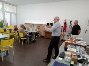 Záber na dlhé stoly s množstvom vystavených kompenzačných pomôcok. Na fotke sú páni z Tyflocompu, ktorí vedú prezentáciu. V pozadí vidieť jeden stôl vôkol ktorého sedia účastníci.